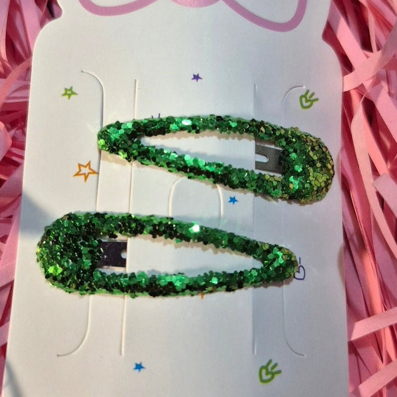 Green glittery hair clip on a white card with a pink bow design, set against a pink shredded paper background.