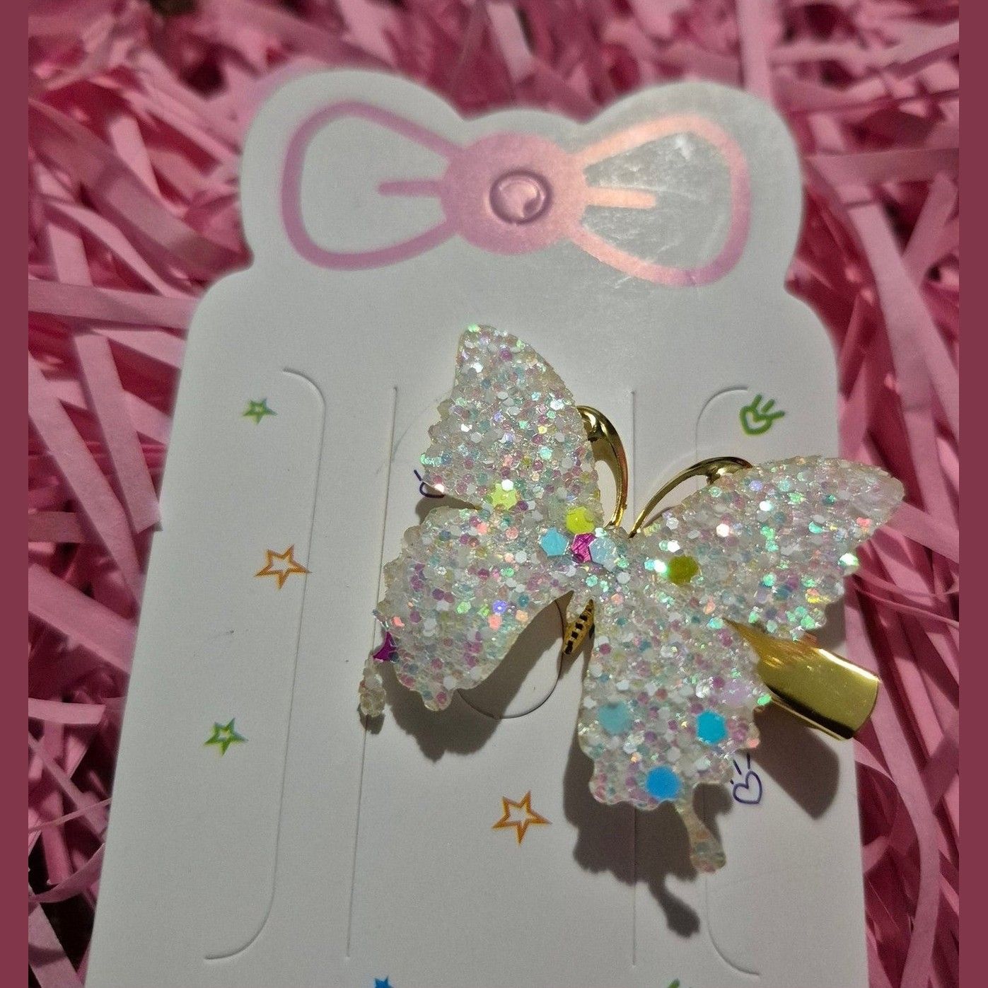 Butterfly-shaped hair clip with glitter on a white card with pink bow design, placed on pink shredded paper.