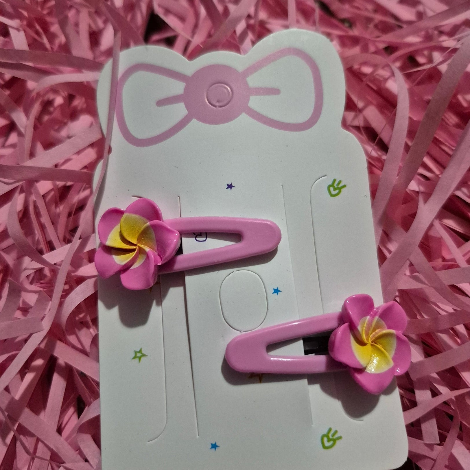 Pink hair clips with flower designs on a white card, placed on pink shredded paper.