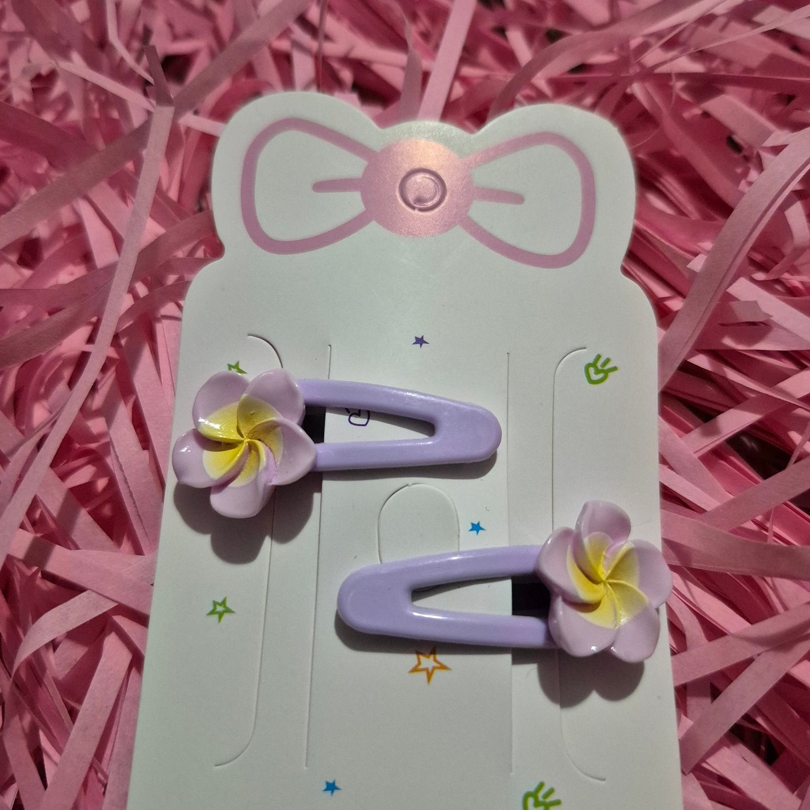 Purple hair clips with floral designs on a pink card with a bow, all set against a pink shredded paper background.
